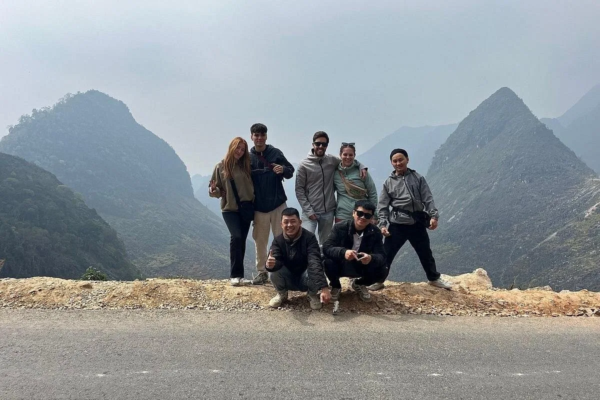A group of travelers pose together on a mountain pass roadside overlooking the layered peaks of the Ha Giang Loop.