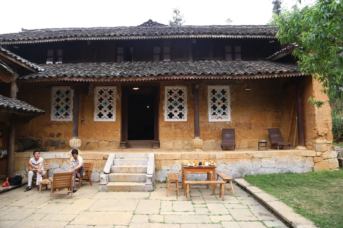 Two men sit and chat outside a traditional earthen house with tiled roof and carved wooden windows in Ha Giang.