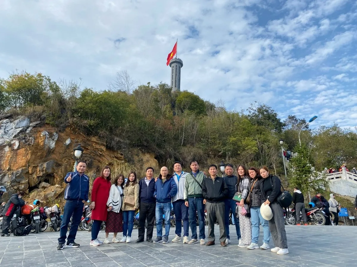 A group of tourists poses on a stone plaza below a rocky hill topped with a flag tower in Ha Giang.