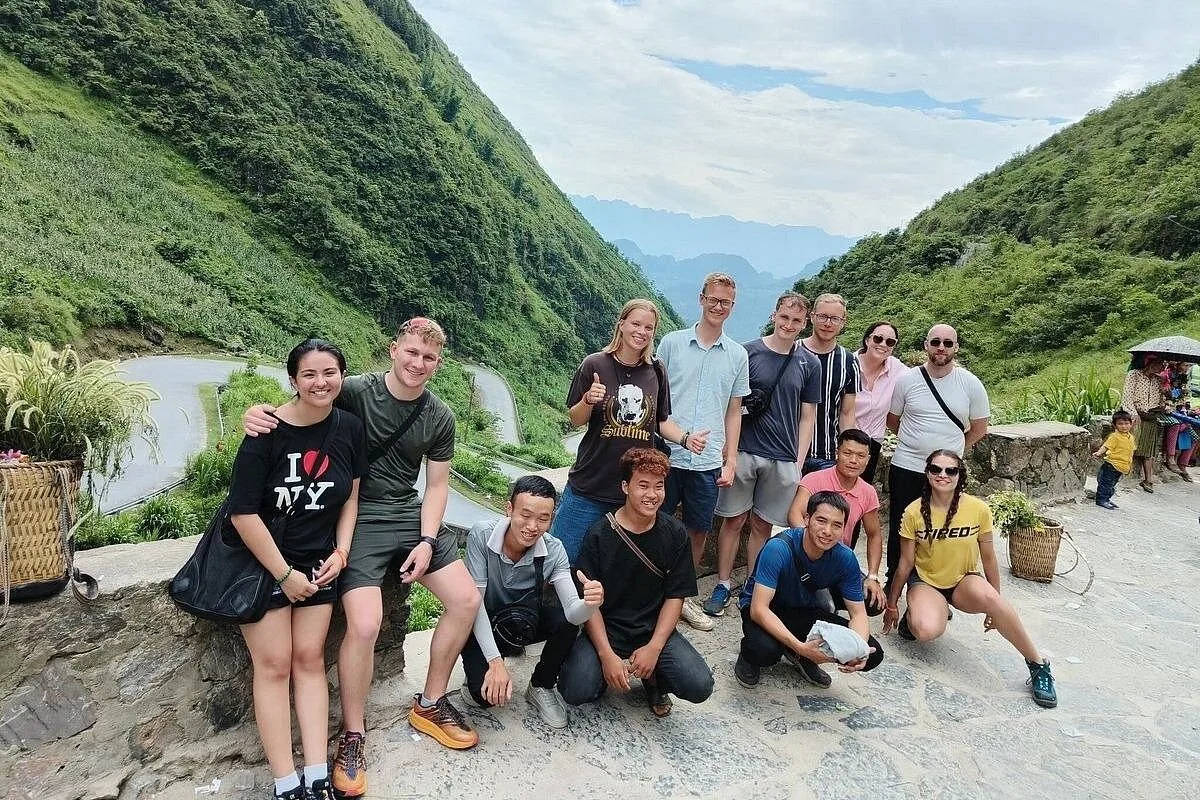 A group of tourists poses together at a scenic overlook above the winding mountain road of the Ha Giang Loop, Vietnam.
