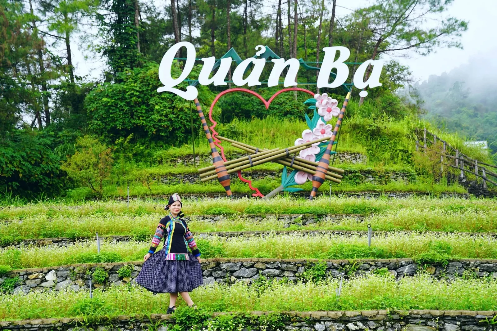 A woman in Hmong ethnic dress poses before terraced flower fields and the Quan Ba decorative sign in Ha Giang.