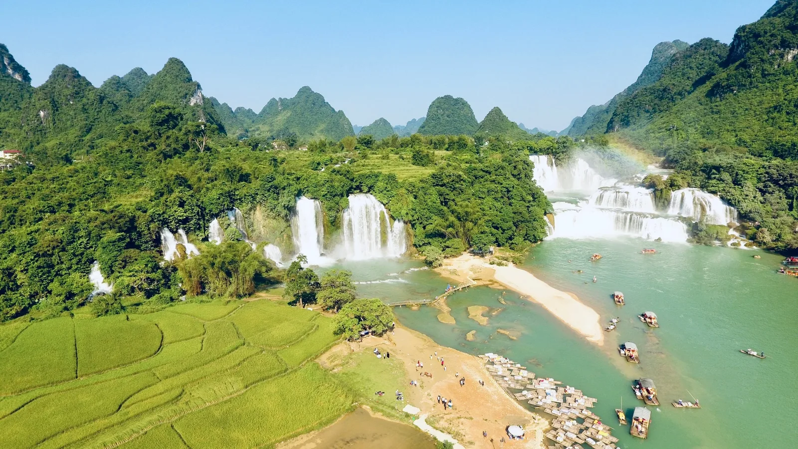Aerial view of Ban Gioc waterfall cascading amid karst mountains, rice fields, and boats on the river in Cao Bang.
