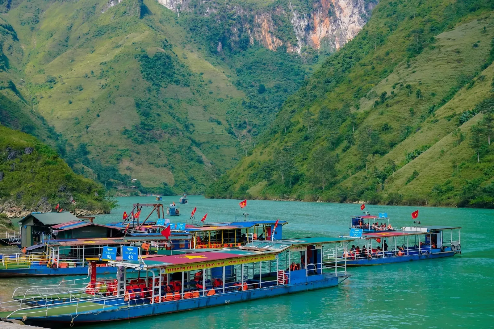 Tourist boats with red flags cruise the turquoise river between steep green mountains along the Ha Giang Loop.