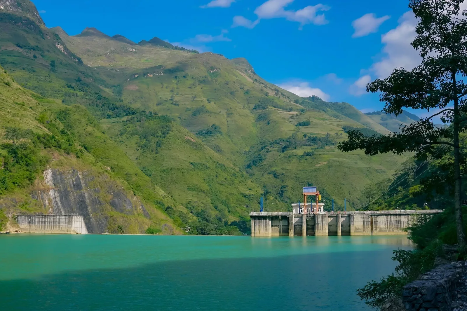 A hydroelectric dam spans a turquoise reservoir surrounded by steep green mountains in Ha Giang.