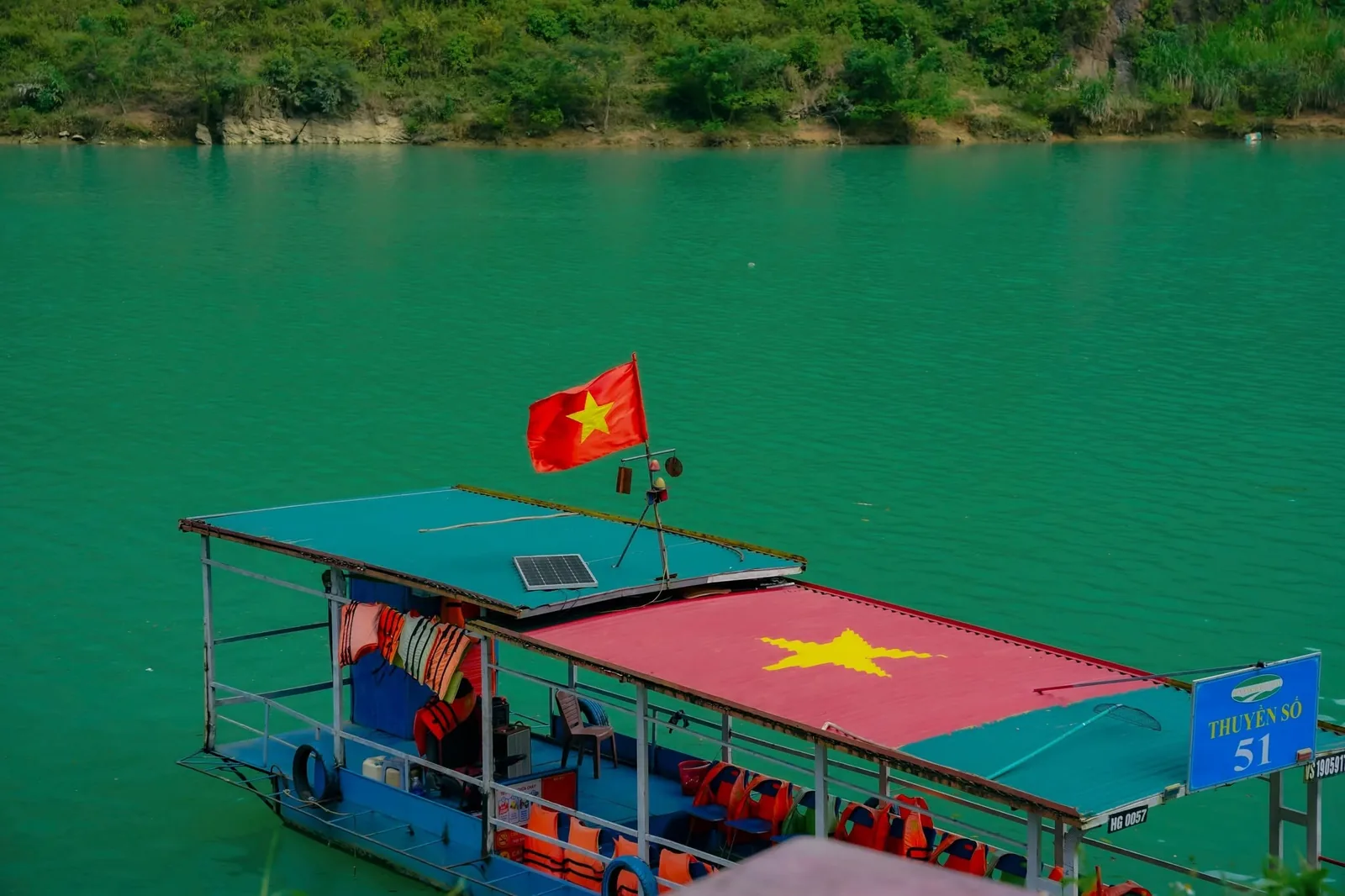 A tour boat with a Vietnamese flag and star-painted roof floats on emerald river water in Ha Giang.