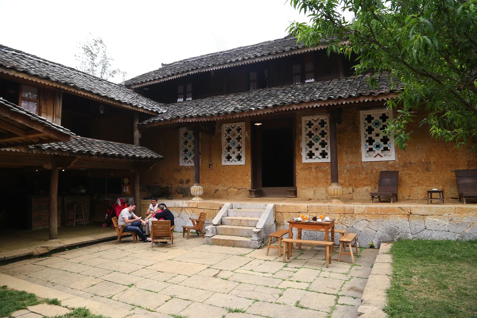 Visitors relax on a stone courtyard in front of a traditional earthen house with tiled roof and carved window shutters in.