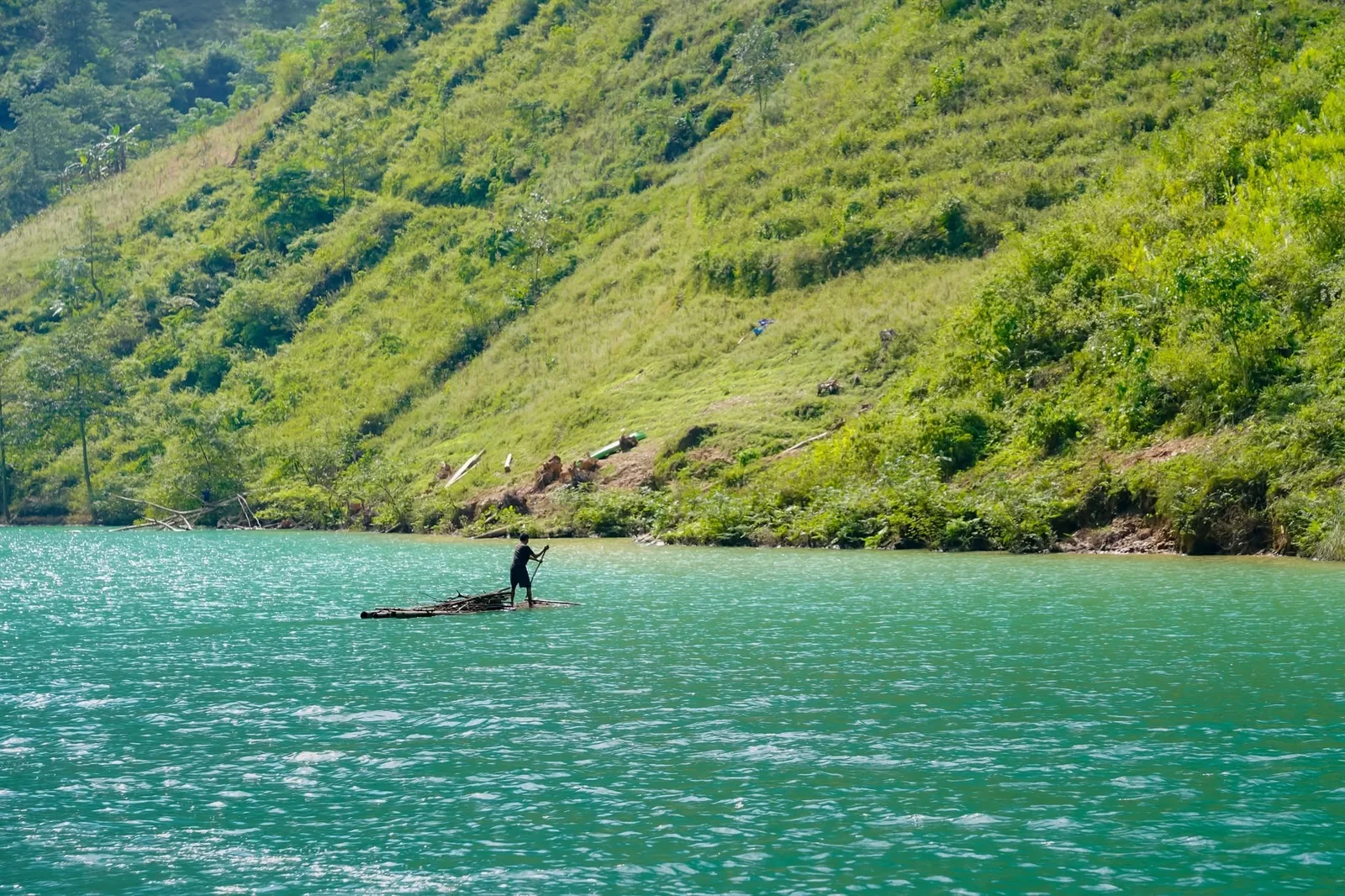 A person poles a small bamboo raft across turquoise water below a steep green hillside in Ha Giang.