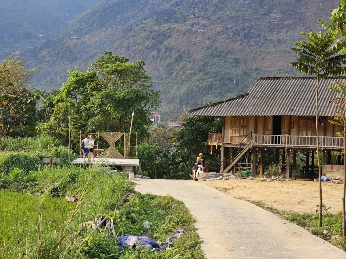 A wooden stilt house sits beside a village road with rice fields and mountains in Ha Giang.